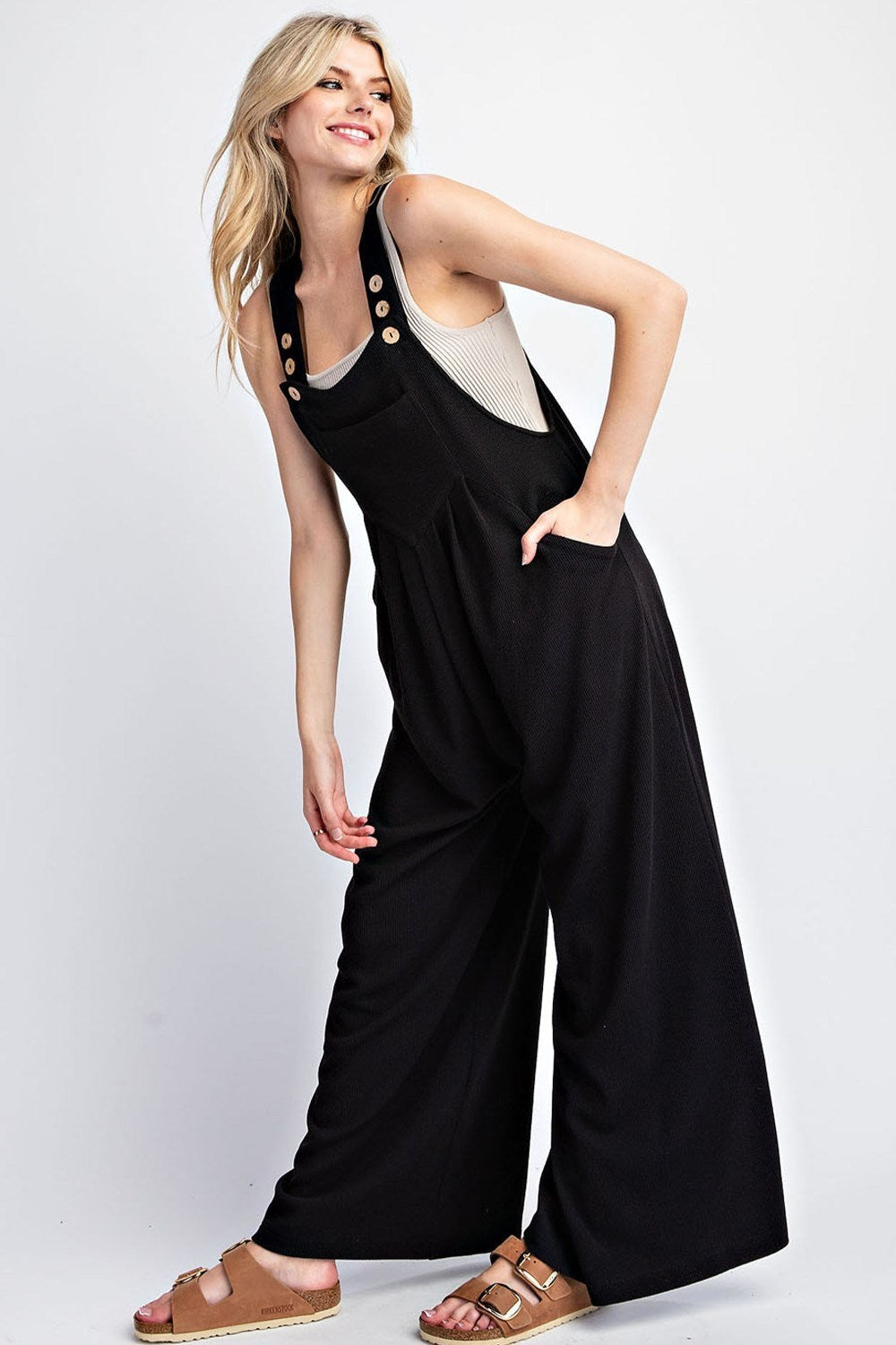 This solid jumpsuit is sleeveless with a square neckline, button adjustable straps, and one front bib pocket. The added pleats provide extra detail, and there are two side hip pockets. It is designed with a wide leg and made of 93% polyester and 7% spandex. This jumpsuit comes in black.