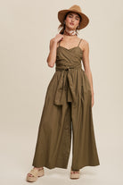 Featuring a removable shoulder strap, this jumpsuit boasts a unique wrap-around design and a flattering smocked body. The wide leg pants are equipped with convenient pockets and a self-tie wrap strap for a customizable fit. Made of 100% cotton, the jumpsuit is finished with a clean hem and is fully lined for added comfort. Color in Deep Olive.