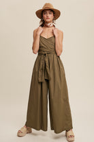 Featuring a removable shoulder strap, this jumpsuit boasts a unique wrap-around design and a flattering smocked body. The wide leg pants are equipped with convenient pockets and a self-tie wrap strap for a customizable fit. Made of 100% cotton, the jumpsuit is finished with a clean hem and is fully lined for added comfort. Color in Deep Olive.