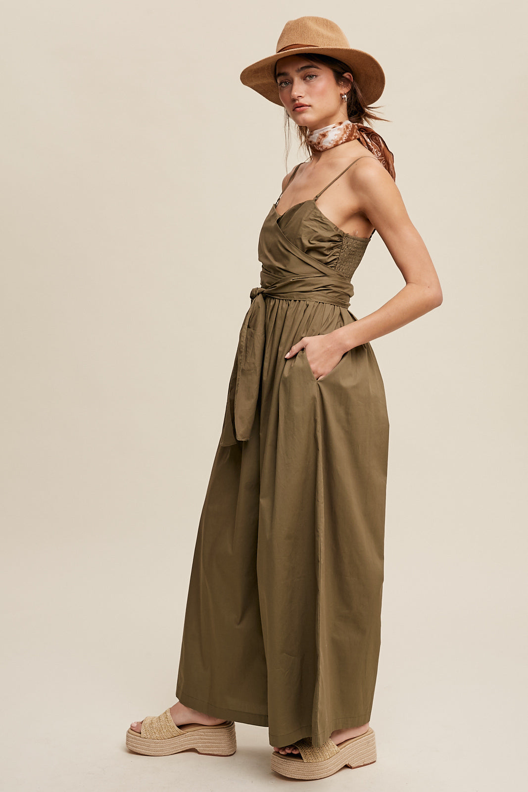 Featuring a removable shoulder strap, this jumpsuit boasts a unique wrap-around design and a flattering smocked body. The wide leg pants are equipped with convenient pockets and a self-tie wrap strap for a customizable fit. Made of 100% cotton, the jumpsuit is finished with a clean hem and is fully lined for added comfort. Color in Deep Olive.