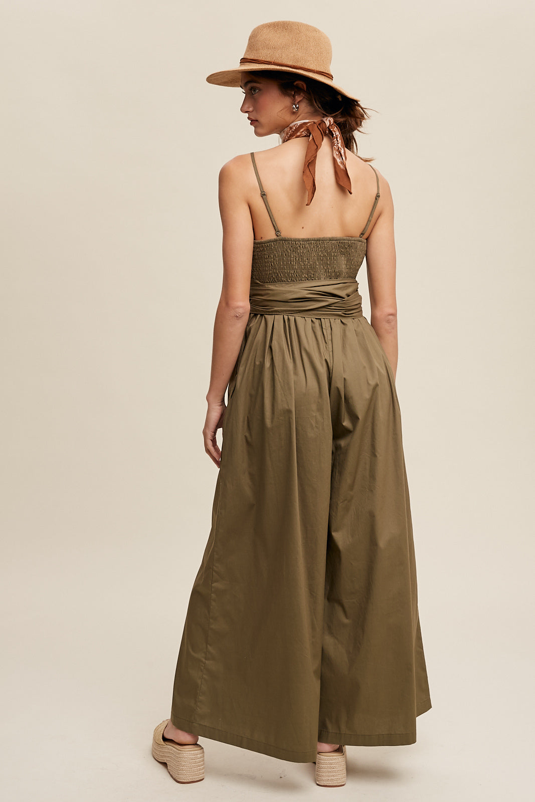 Featuring a removable shoulder strap, this jumpsuit boasts a unique wrap-around design and a flattering smocked body. The wide leg pants are equipped with convenient pockets and a self-tie wrap strap for a customizable fit. Made of 100% cotton, the jumpsuit is finished with a clean hem and is fully lined for added comfort. Color in Deep Olive.