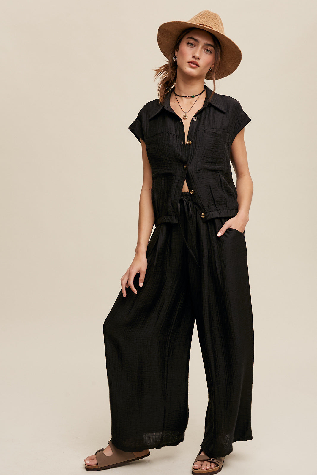 Consisting of a button down top and pleated wide leg pants, this set features a collared neck, short sleeves, and an elastic hem. The pants are designed with wide legs, pockets on the side seam, an elasticized drawstring waist with a tie string, and a clean hem. Made with 45% Rayon, 37% Tencel, and 18% Nylon, this set is available in black.
