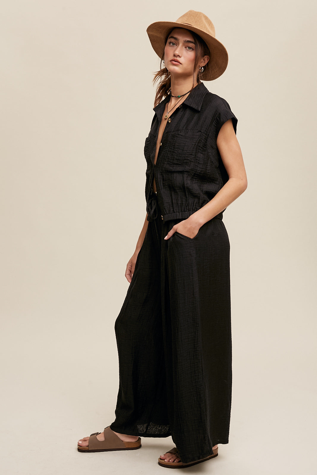 Consisting of a button down top and pleated wide leg pants, this set features a collared neck, short sleeves, and an elastic hem. The pants are designed with wide legs, pockets on the side seam, an elasticized drawstring waist with a tie string, and a clean hem. Made with 45% Rayon, 37% Tencel, and 18% Nylon, this set is available in black.