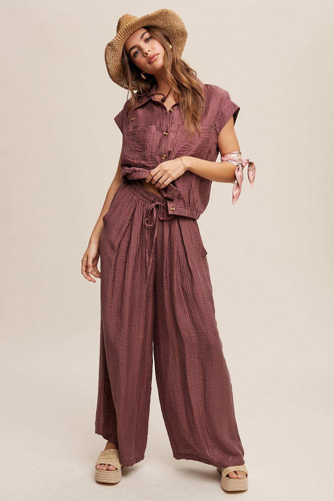 Consisting of a button down top and pleated wide leg pants, this set features a collared neck, short sleeves, and an elastic hem. The pants are designed with wide legs, pockets on the side seam, an elasticized drawstring waist with a tie string, and a clean hem. Made with 45% Rayon, 37% Tencel, and 18% Nylon, this set is available in Red Bean.