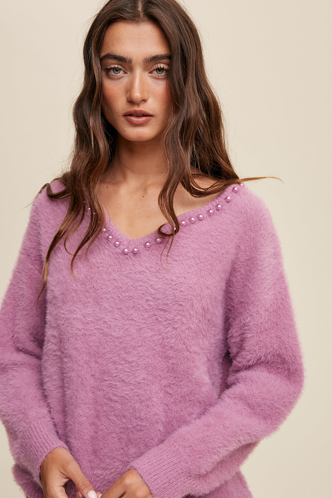 The Deep V-Neck Peal Detail Fuzzy Knit Sweater is composed of a soft touch Fuzzy Knit material, featuring a deep V-neckline and delicate pearl detailing. It boasts long sleeves and is finished with ribbed accents on the neck, cuffs and hem. Made of 65% acrylic and 35% polyester, this sweater is available in a lovely Lilac hue.